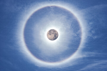 rare natural phenomenon created when sunlight hits ice crystals suspended high in the atmosphere, circular rainbow halo around the moon among blue sky and white clouds