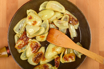 An overhead shot shows light green bell pepper slices browning in a dark frying pan, with a wooden spatula resting among them on a wooden surface