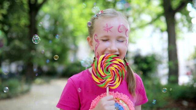 Adorable happy little girl with princess makeup and colorful lollipop having fun in the park