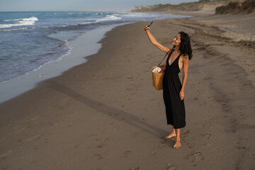 Brunette Woman in Black Dress Taking a Selfie on the Beach with Straw Bag