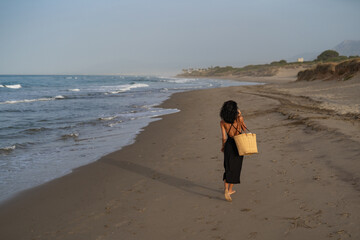 Brunette Woman in Black Dress Walking and Talking on Phone at the Beach with Straw Bag