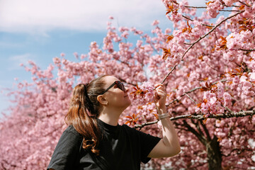Young woman in sunglasses gently smelling cherry blossoms. Perfect for nature campaigns, springtime wellness ads, beauty editorials, or seasonal branding visuals.