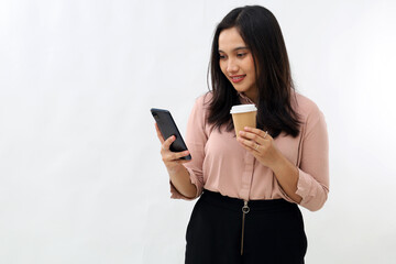 Naklejka premium Portrait of modern Asian woman looking at mobile phone, holding coffee paper cup, using smartphone app, standing on white background.