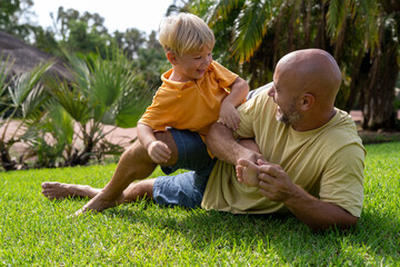 family and love concept - young father tickling his little son in park. dad tickles his son's legs on the grass. dad with little son having fun on the green lawn