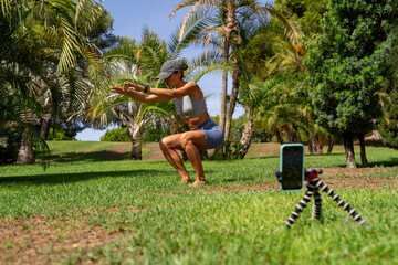 Athletic woman blogger in sportswear shoots video on camera as she does exercises in park. Sport and recreation concept. Healthy lifestyle.