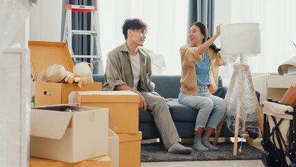 Young happy Asian couple help each other moving box full of stuff unpacking furniture on sofa couch in living room at new house. Husband and wife move new house, Apartment Rent And Ownership Concept.