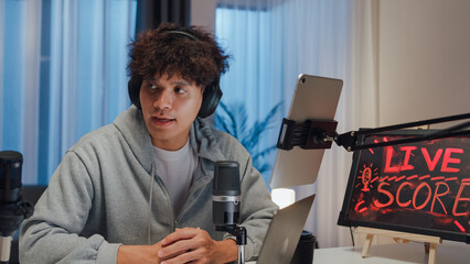 Young happy Asian couple wear headphone sit on desk online live streaming football live score excited cheering watch soccer sport game on laptop talk with audience in small studio at home at night.