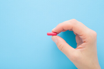 Young adult woman hand fingers holding and showing red pill for improve of iron level in blood on light blue table background. Pastel color. Closeup. Top down view.
