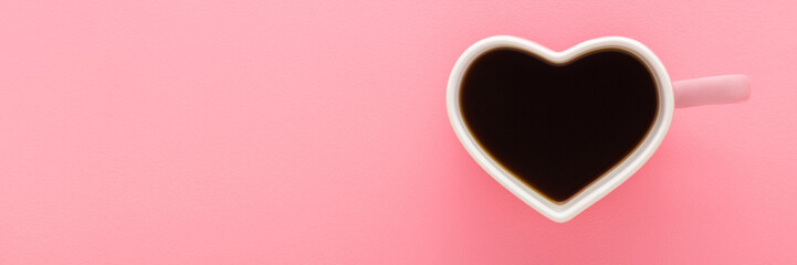 Heart shape white cup with fresh black coffee on light pink table background. Pastel color. Wide banner. Closeup. Empty place for text. Top down view.