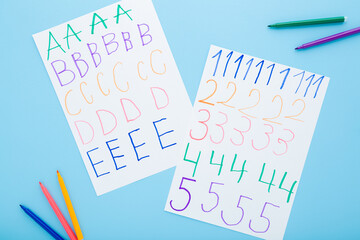 Colorful markers and written different english letters and numbers on white paper sheets on light blue table background. Pastel color. Closeup. School exercises for little child. Top down view.