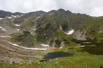 Mountain lake nestled in alpine valley with snow patches on rocky peaks and green meadows under cloudy sky