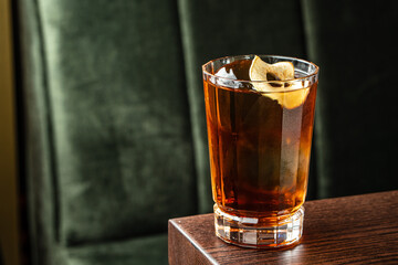Dark amber cocktail with ice and dried fruit garnish served in a faceted glass on a wooden table with a green velvet background for a cozy lounge atmosphere