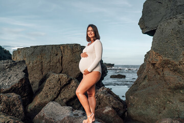 Pregnant woman standing among rocks by ocean, hand on belly, embodying stability, courage, and...