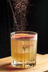 Golden cocktail in a faceted glass garnished with citrus slice and vibrant powder being sprinkled from above against a dramatic dark background