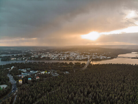 A dramatic sunset over a northern city, with sunrays piercing through storm clouds and illuminating the urban and forested landscape. A peaceful river reflects the golden light.