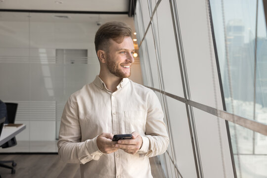 Businessman, employee dressed in casual shirt distracted from smartphone usage, look out window with optimistic expression, wait for business call, feel satisfied with productive workday using tech