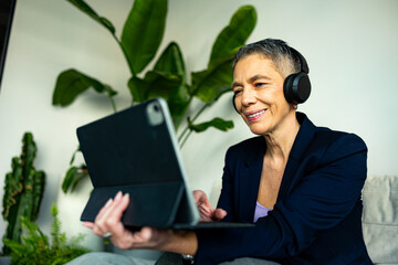 Happy mature businesswoman attending a video conference on a tablet while wearing headphones and working remotely from home