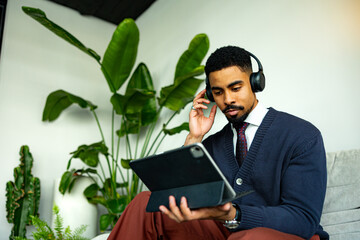 Young businessman wearing headphones using tablet and working remotely surrounded by plants in a modern office