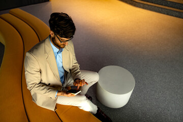 Businessman sitting comfortably on a modern sofa in an office lobby, using a smartphone for communication and browsing online