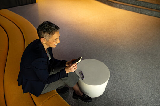 Smiling businesswoman sitting comfortably in a modern waiting area, engaging with her smartphone while enjoying a moment of relaxation
