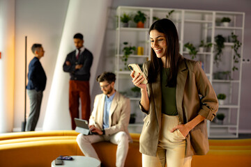 Businesswoman smiling and using mobile phone in modern office with colleagues working in background