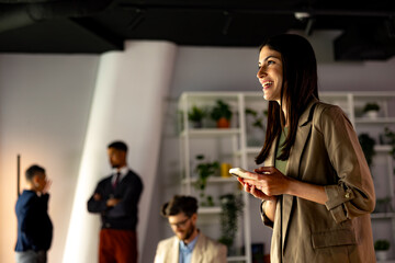 Businesswoman using mobile phone and smiling while her colleagues are standing in the background in a modern office