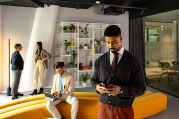 Young businessman texting on his phone while his colleagues work in the background in a modern office