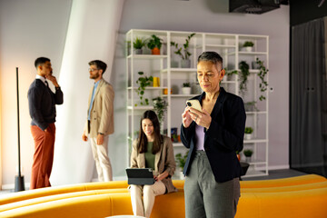 Mature businesswoman using mobile phone in modern office environment, with colleagues discussing and working in the background