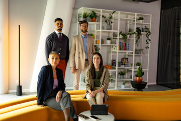 Four businesspeople posing confidently in a modern office, surrounded by greenery and featuring a stylish yellow sofa, embodying teamwork