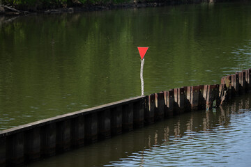 Red triangular navigation sign on a wooden barrier in a calm green river. Ideal for use in travel, boating safety, river management, or minimalist nature themes.