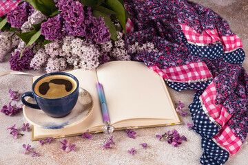 Spring flatlay with an open notebook, a bouquet of blooming lilac flowers, a cup of coffee, a pen, a purple shawl on a light background.