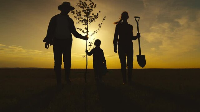 A family of three is walking through a field with a tree in the foreground. The man is holding a shovel and the woman is holding a tree sapling