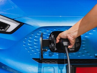 A close-up image showcases an electric vehicle charging at a public charging station. The sleek white car, with its futuristic headlights, is connected to a charging cable with a glowing green plug.