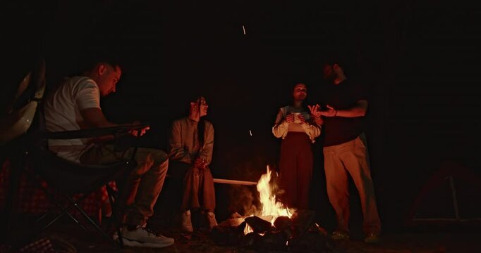 Friends gather around a bonfire at night, sharing stories and laughter. The warm firelight illuminates their faces as they enjoy each other's company.