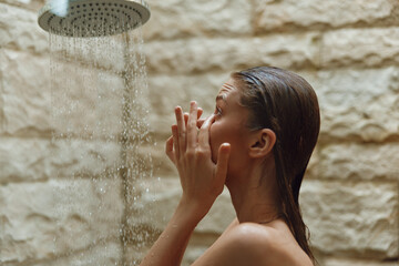 Young woman taking a refreshing shower with water falling, natural stone wall background, wet hair, relaxed mood, personal care moment, indoor bathroom setting.