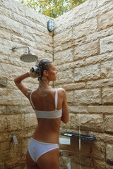 young woman in white bikini washing hair under outdoor stone shower with natural light and green trees in background, summer wellness and skincare moment