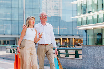Senior couple shopping downtown, carrying colorful bags, smiling and walking together while...
