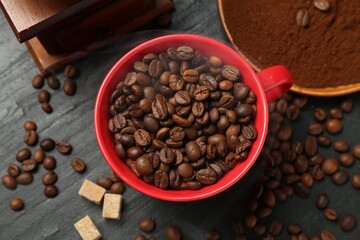 Aromatic coffee beans in cup, grinder and brown sugar on black table, flat lay