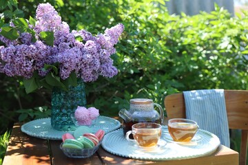Beautiful lilac flowers in vase, sweets and tea on wooden table in garden