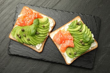 Delicious sandwiches with avocado, salmon, cream cheese, basil and capers on black textured table, top view
