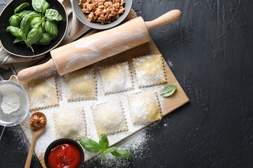 Uncooked ravioli, rolling pin, bolognese, sauce and basil on black table, flat lay