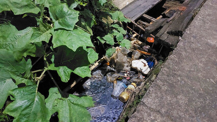 An outdoor scene shows a dirty drainage ditch filled with trash and stagnant water, partially...