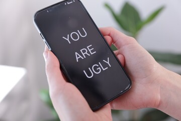 Cyber bullying. Woman holding smartphone with text You Are Ugly on blurred background, closeup