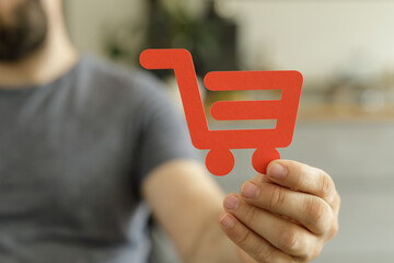 Close-up of a man's hand holding a red paper shopping cart icon.  E-commerce, online shopping...