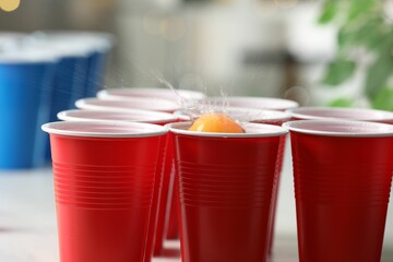 Orange ball falling into plastic cup on light table against blurred lights, closeup. Beer pong game