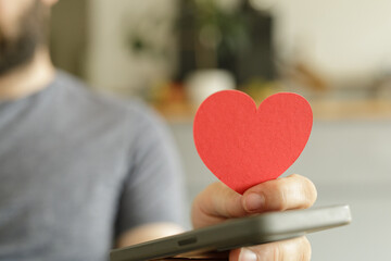Close-up of a man's hand holding a red paper heart shape near his phone, symbolizing love,...