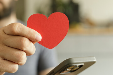 Close-up of a man's hand holding a red heart shape, near a smartphone.  Shows love, care,...