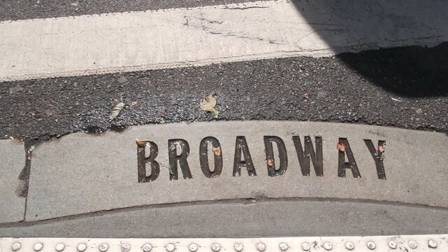Broadway street sign, people on pedestrian zebra crossroad. New York City Manhattan road intersection. Text on asphalt near crosswalk, shadows on crossroad ped crossing. Traffic signage, United States