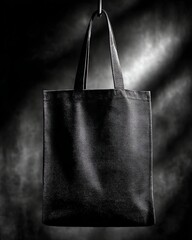 A plain black tote bag with two handles is hanging against a dark, dramatic background with soft lighting
