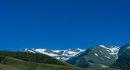 The photo shows picturesque mountains with green slopes and snow patches on the peaks. A bright...